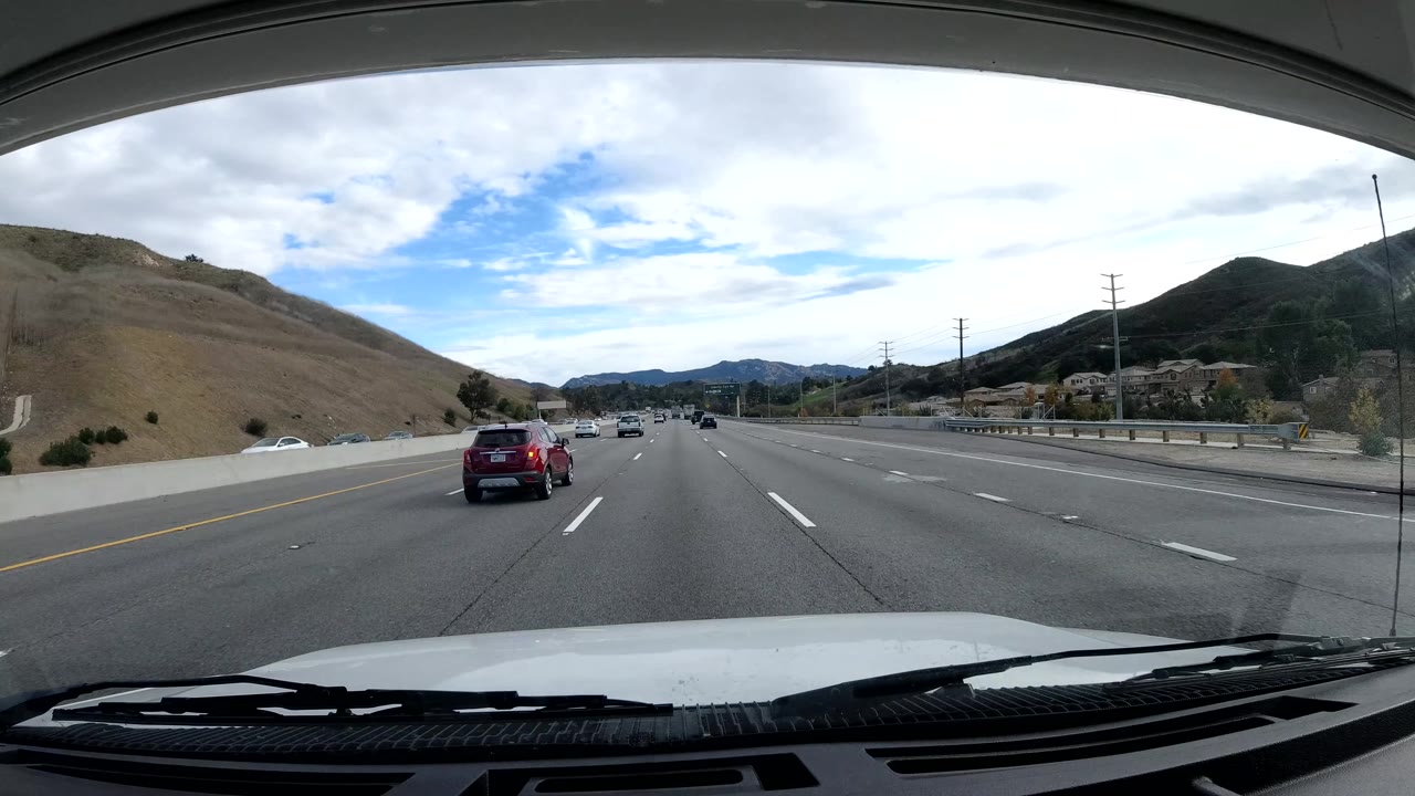 Highway lanes with a red SUV ahead, brown hillside, guardrail, and distant mountains.