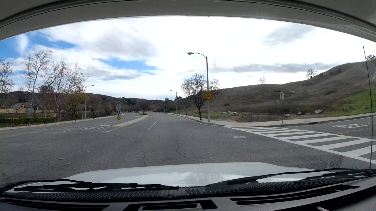 Malibu dashcam view at a quiet road junction with white crosswalk markings, open hillside, sparse trees, road signs, and no clearly visible people.