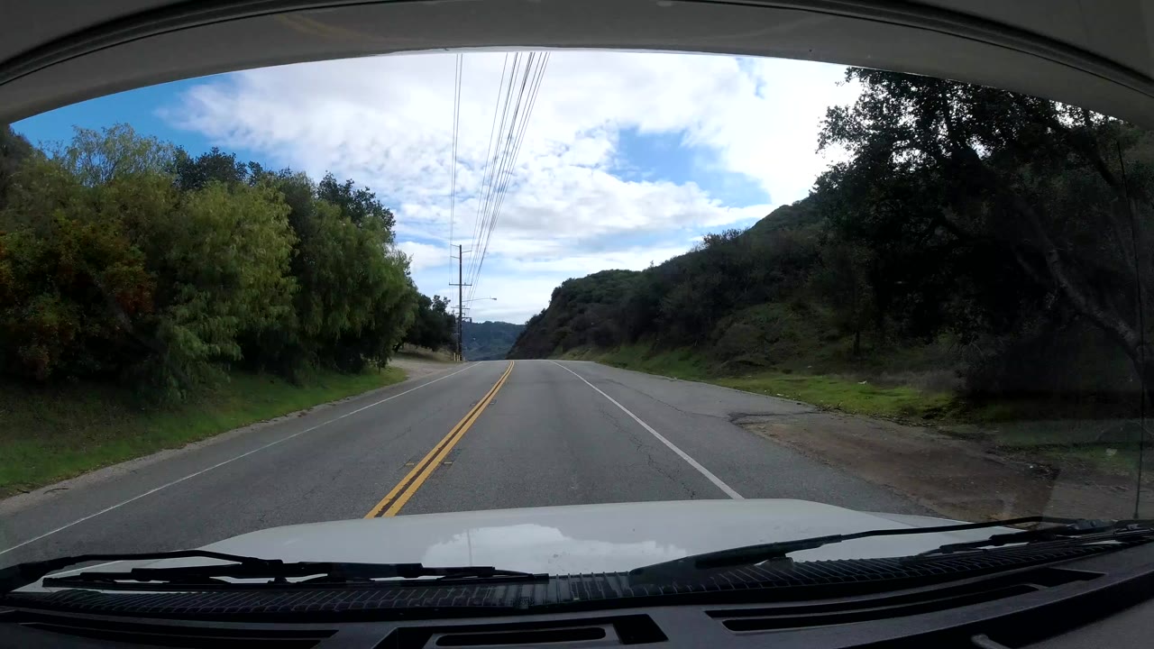 In-vehicle view along a Malibu, California road with lane markings, dense trees at the roadside, overhead utility lines, a bright cloudy sky, and no visible people.
