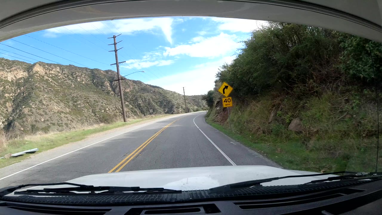 Dashcam-style view in Malibu, California, looking down a curving two-lane road with a yellow curve warning sign, utility poles, hillside brush, blue sky, and no visible people.
