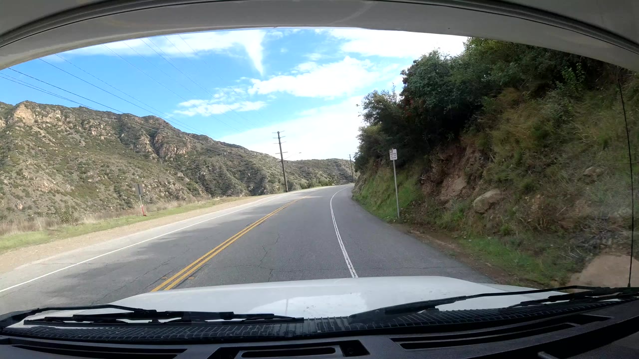 Two-lane hillside road continues past brush and utility lines.