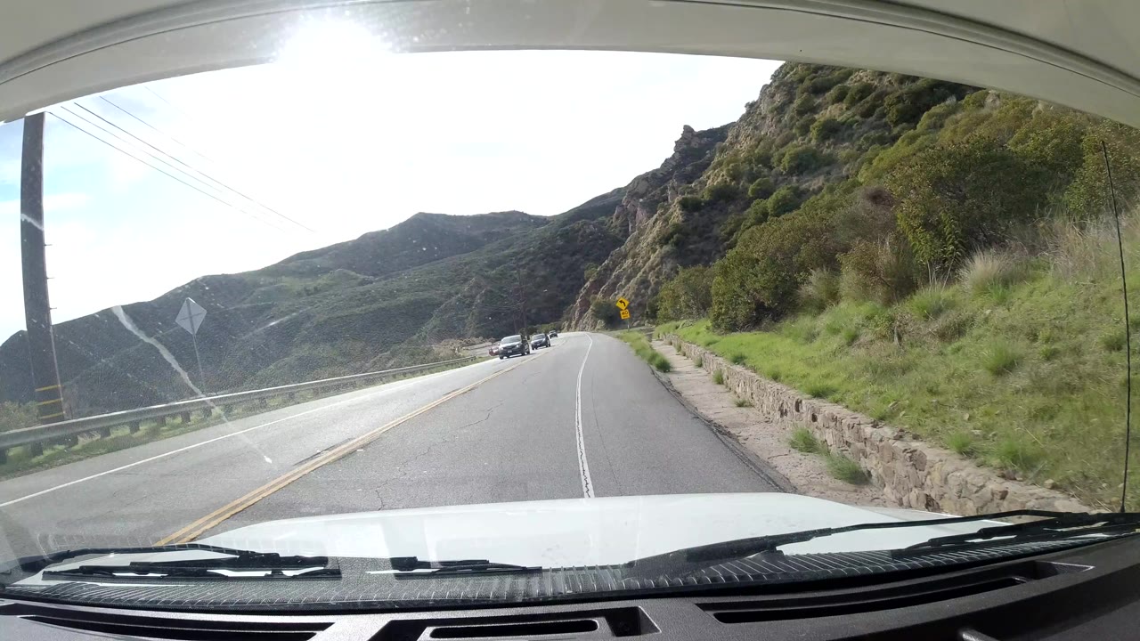 Malibu, California road scene from inside a vehicle, showing a right-hand curve, roadside guardrail, hillside slopes, several oncoming cars, glare on the windshield, and no visible people.