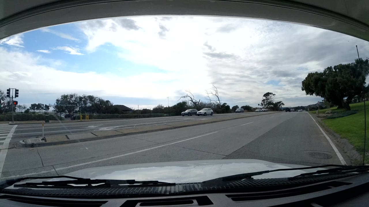 Malibu windshield view at the start of the clip, showing a broad paved road, passing cars, green roadside grass, trees, cloudy daylight, and no visible people.