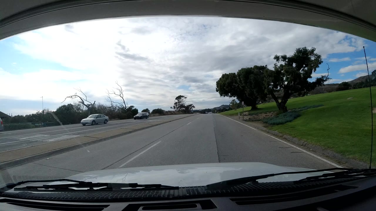 Malibu road view one second in, with the vehicle hood in the foreground, a curving lane ahead, cars to the left, a green hillside, large roadside trees, and no visible people.
