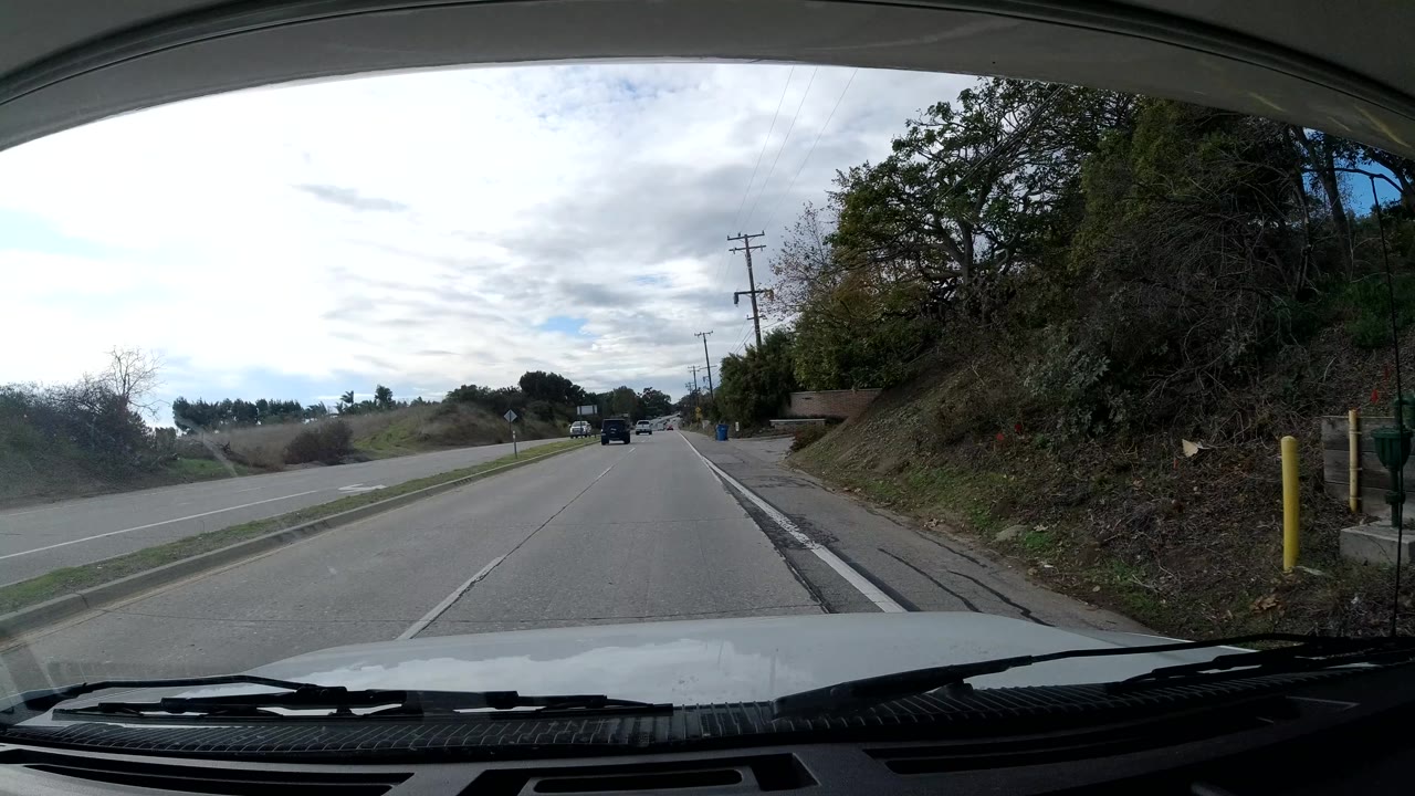 Malibu windshield view with the road stretching ahead, hillside brush and trees on the right, utility poles along the roadside, cars in the lane distance, and no visible people.