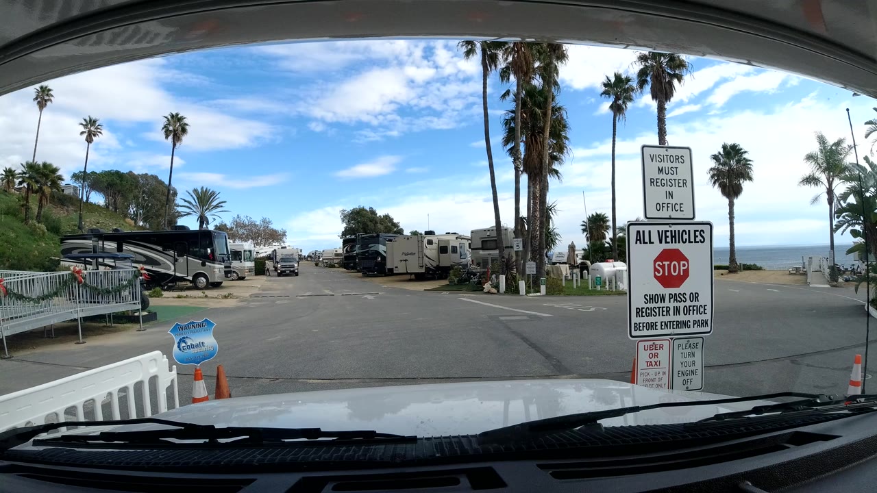 Malibu, California parking entrance seen through a vehicle windshield, with RVs to the left, signs on the right, palm trees, ocean horizon, bright clouds, and no visible people.