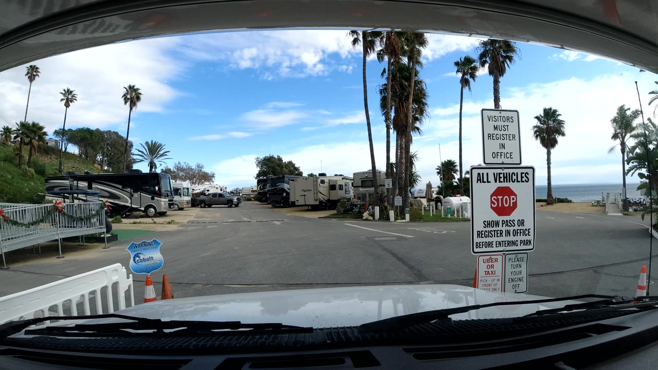 Windshield-level view in Malibu, California approaching a campground entrance sign, with parked RVs, tall palms, a bright sky, a distant ocean view, and no visible people.