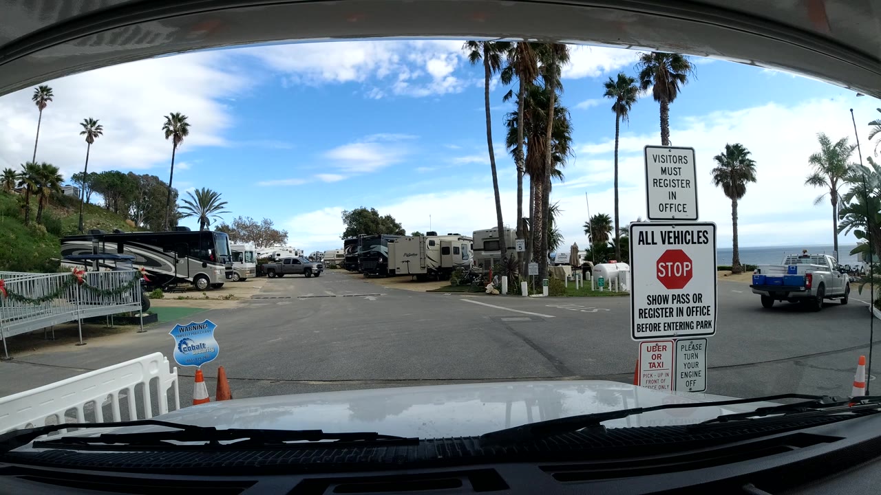 Vehicle POV in Malibu passing the campground sign area, with a blue pickup at right, RVs and palms ahead, ocean visible beyond the road, and no visible people.