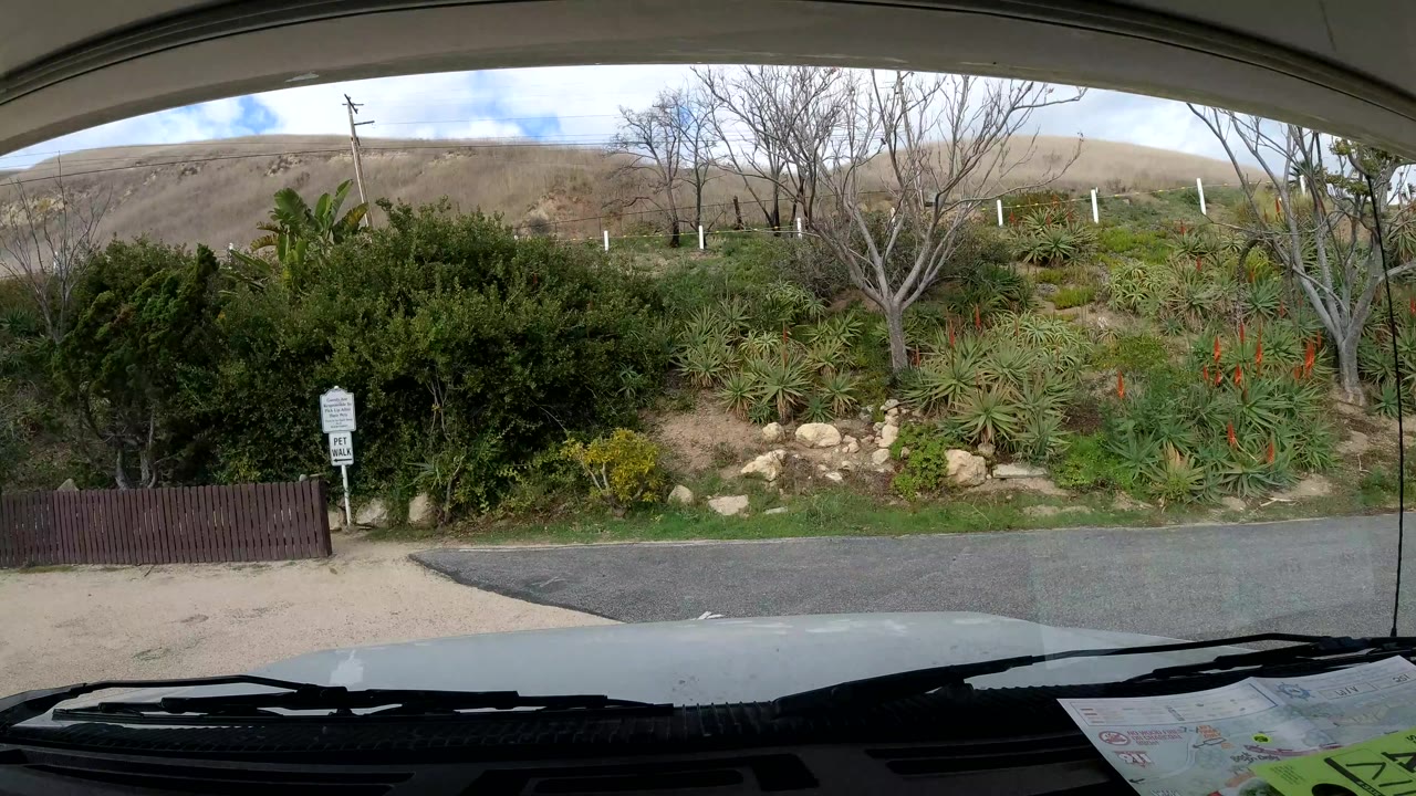 Malibu, California windshield view with dense shrubs, rocks, leafless trees, a low hillside, a small roadside sign, and no visible people.