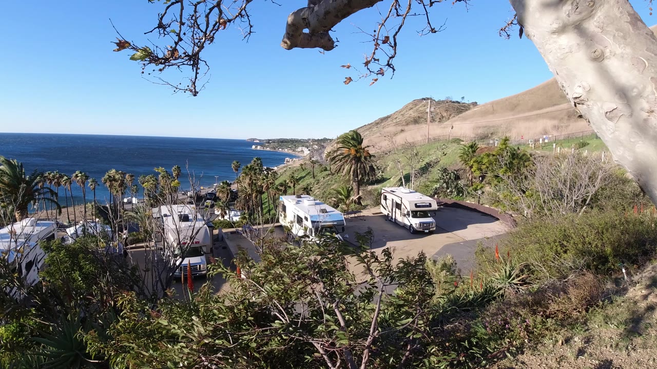 Malibu coastal park scene with two prominent RVs on a paved road, palms below, dry hillside to the right, and no visible people.