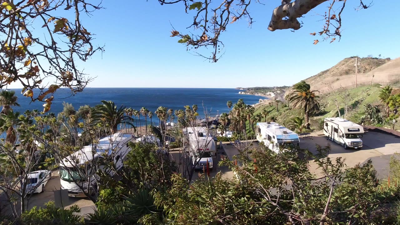 Elevated Malibu coastal view at the start of the clip, with dry branches overhead, parked RVs and cars below, palm trees, hillside, blue ocean, and no visible people.