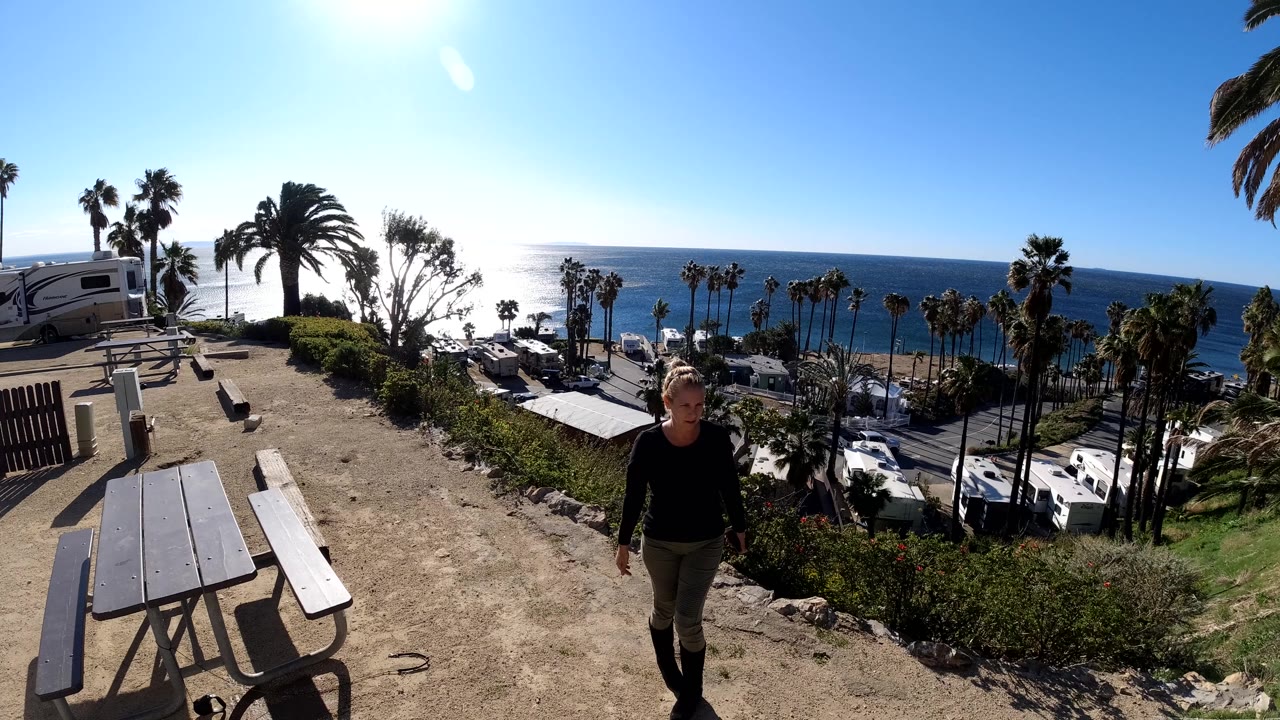 A woman walks toward the camera near picnic tables at a Malibu overlook, with palm trees, parked RVs, ocean water, and bright sun glare in the background.
