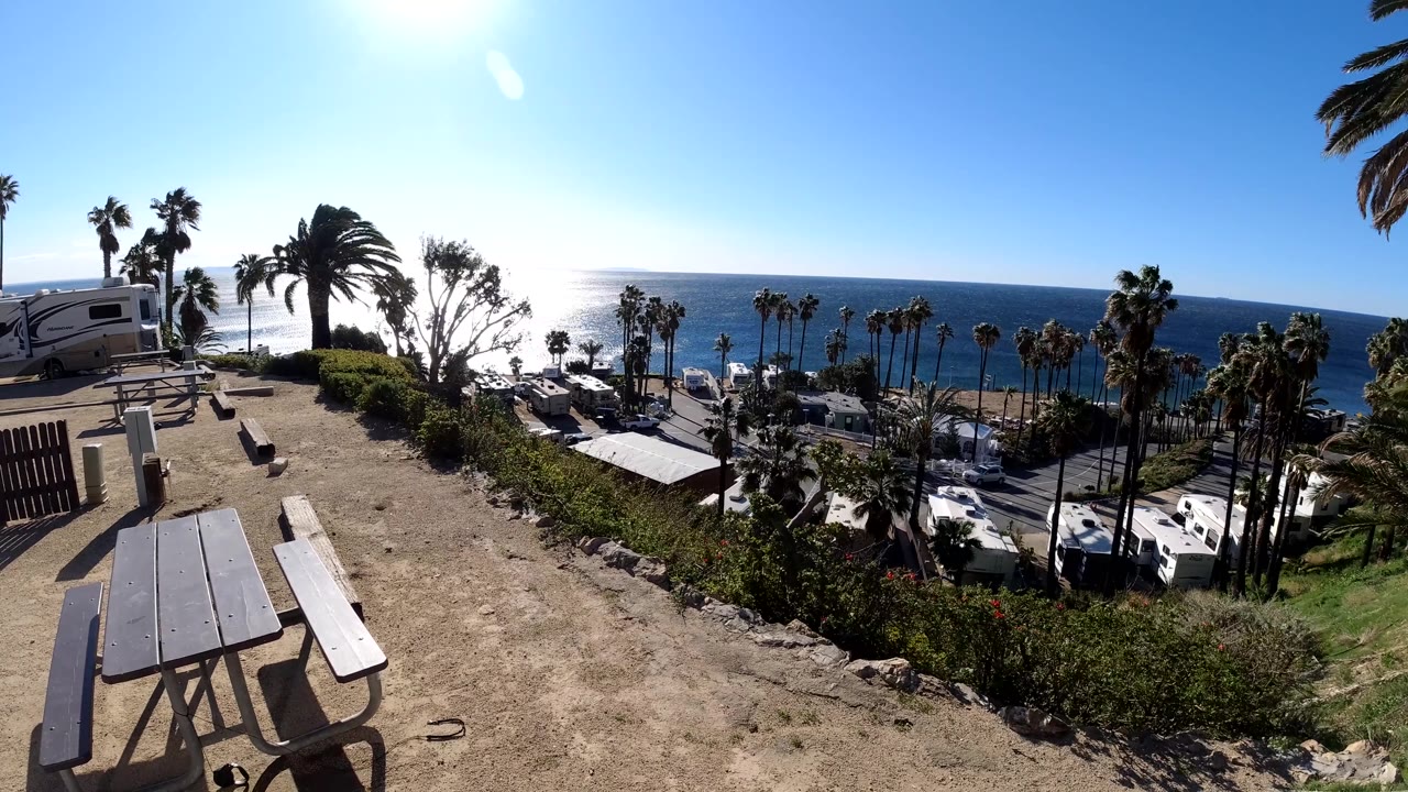 Empty picnic tables sit on the dirt overlook in Malibu, with palm trees, parked RVs, shrubs, and the blue ocean visible under strong midday light, no visible people.