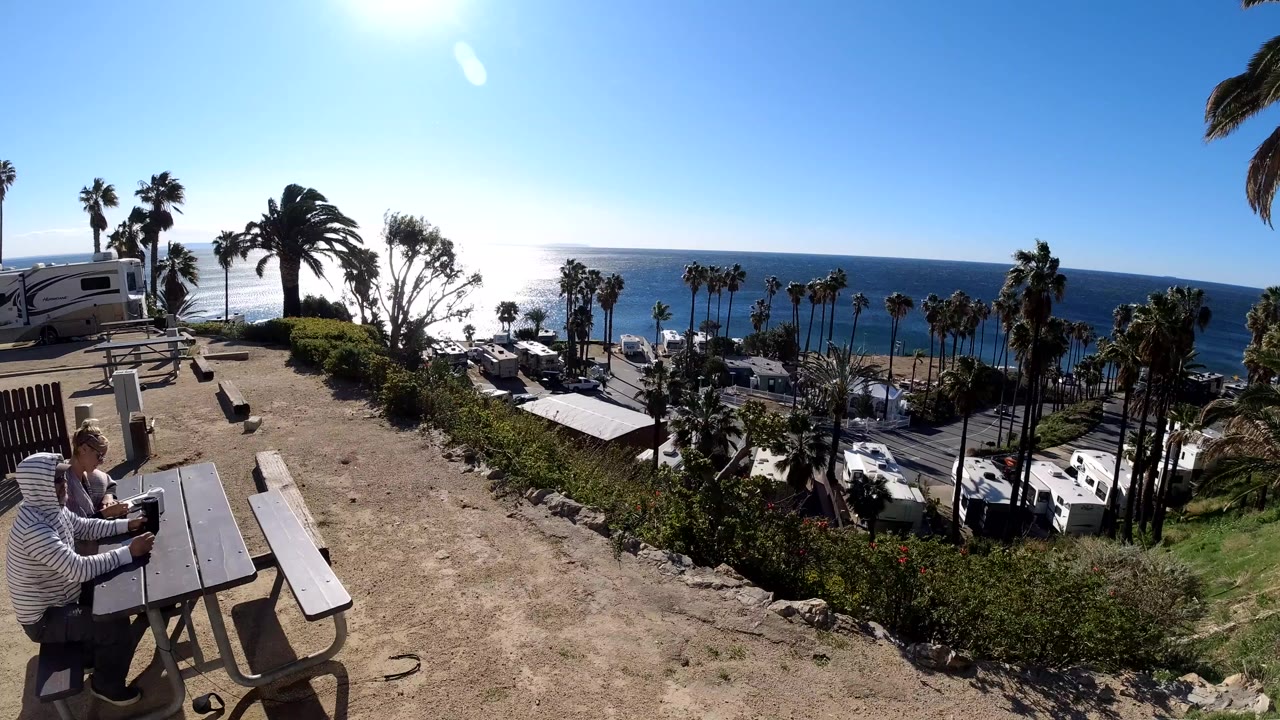 At the start of the Malibu clip, two people sit at a picnic table in the lower left while sun glare reflects off the ocean beyond palm trees and parked vehicles.