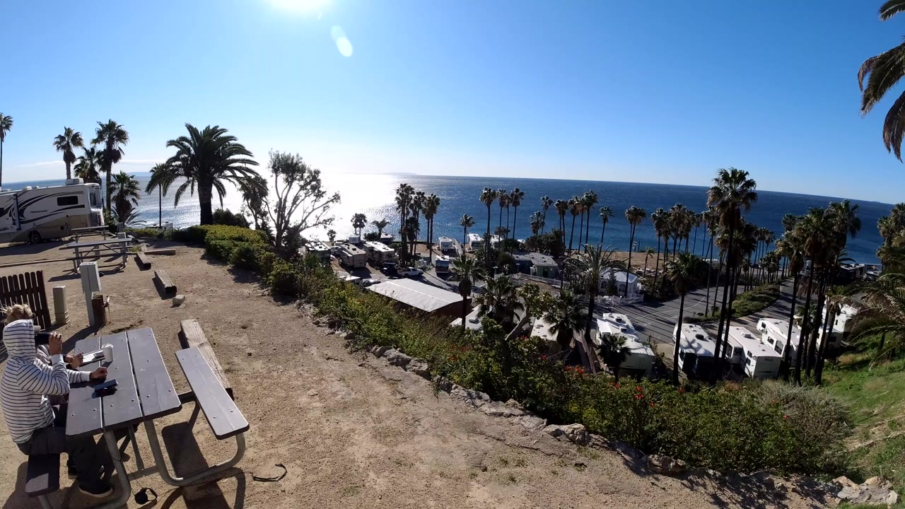 Wide Malibu overlook frame with picnic tables at left, parked RVs below, palm trees across the slope, and bright sun glare over the ocean.