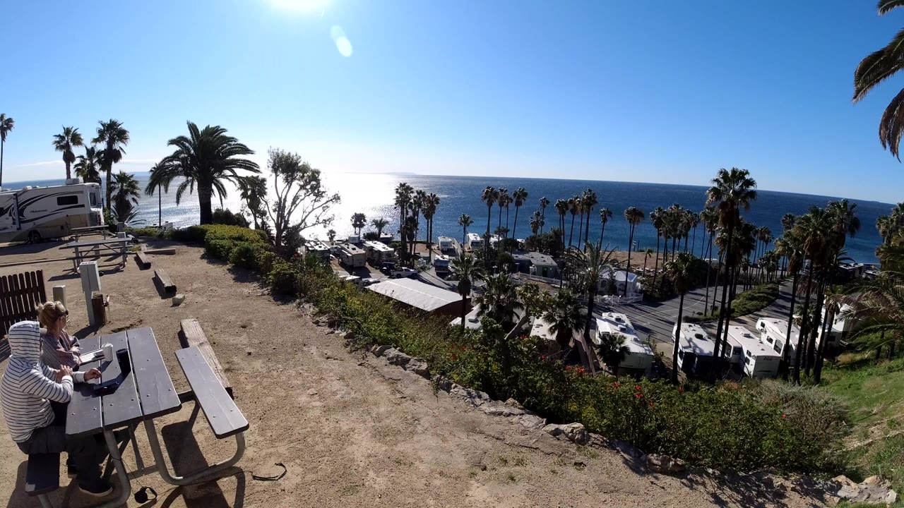 Two people near picnic tables above RVs and the ocean.