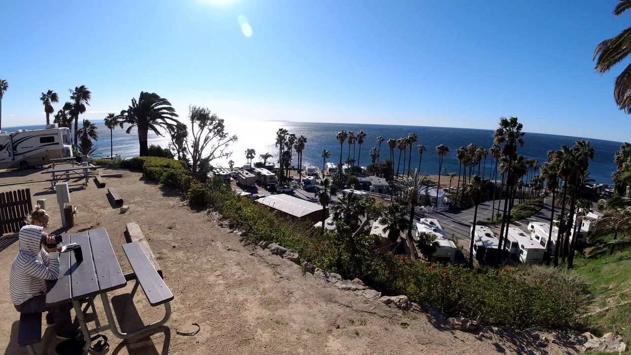 A person in a striped hooded top holds a camera beside a picnic table on a Malibu hillside overlook, with palm trees, parked vehicles, rooftops, and ocean in the background.