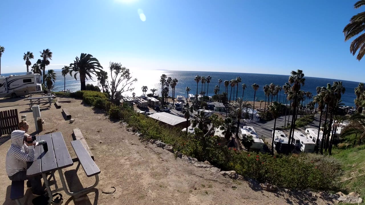 The person remains near the picnic table on the Malibu overlook while the bright ocean horizon, palm trees, RVs, and hillside vegetation fill the view below.