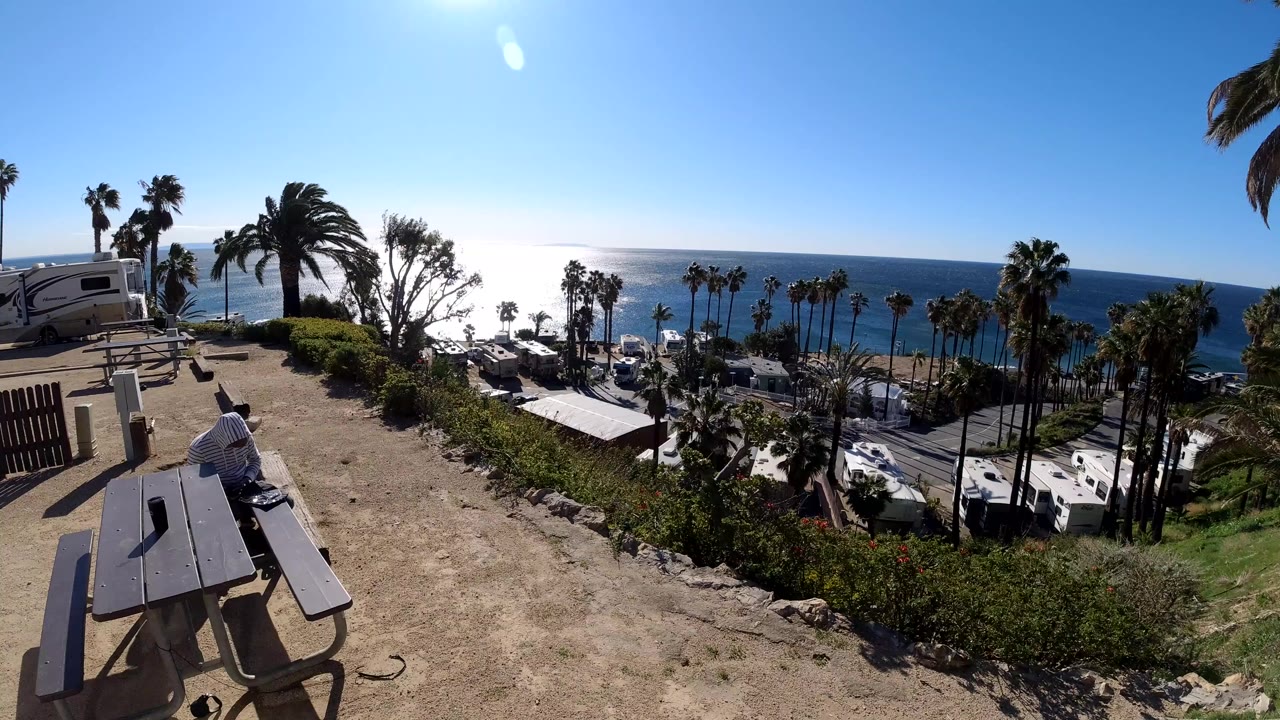 Malibu coastal overlook with a picnic table in the foreground, palms and parked vehicles below, blue ocean on the horizon, and no visible people in this opening frame.