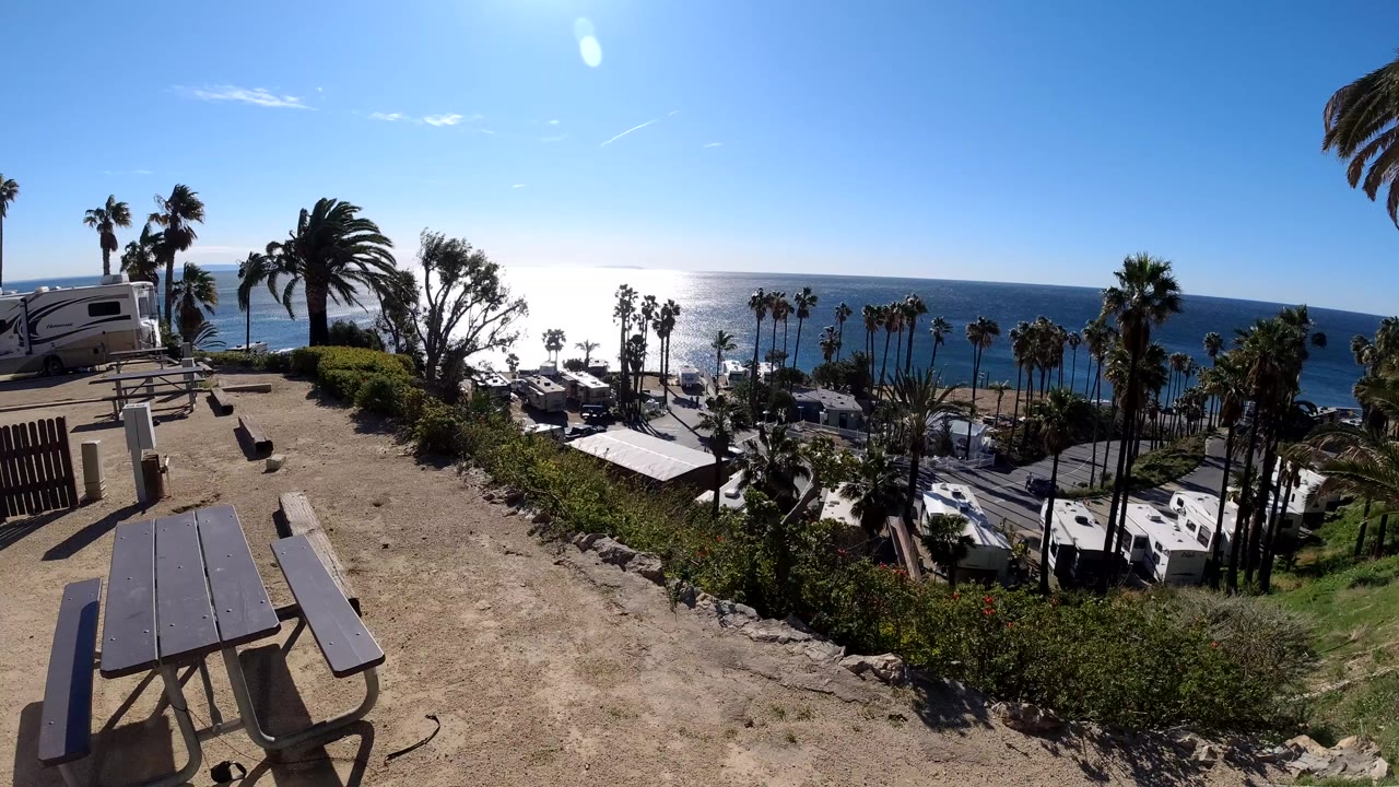 Malibu coastline frame with sunlit ocean on the horizon, clustered RVs in the midground, palms along the slope, picnic tables at left, and no visible people.