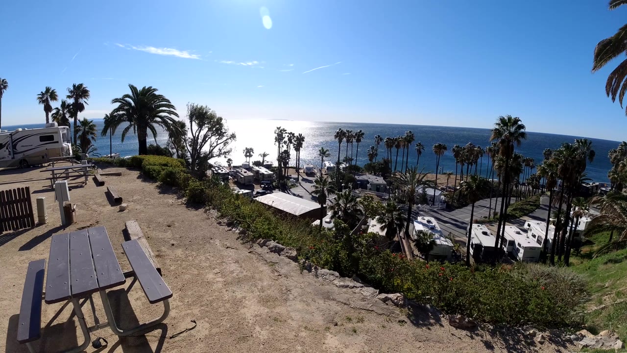 Wide Malibu coastal landscape with empty picnic tables in front, shrubs along the drop, RVs and palm trees below, bright water beyond, and no visible people.