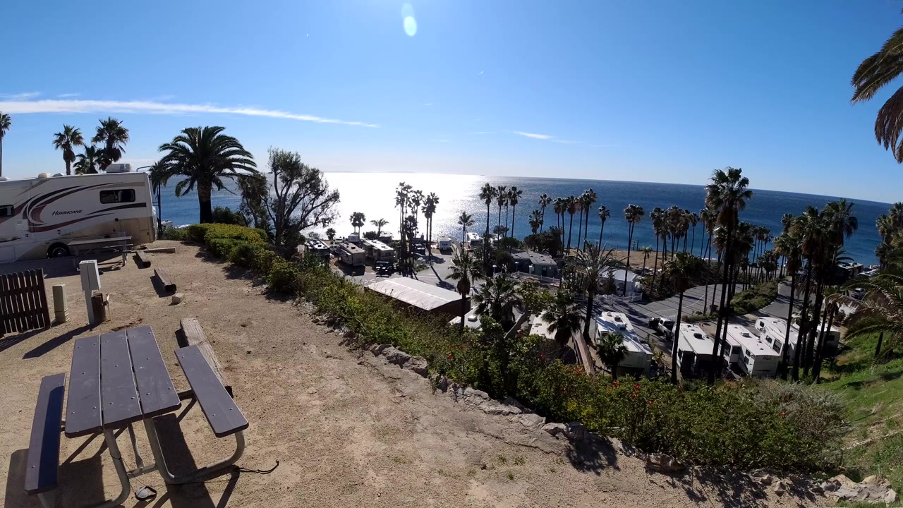 Wide Malibu coastal overlook with a picnic table in the foreground, an RV parked at left, palm trees below, and the ocean horizon in bright sun with no visible people.