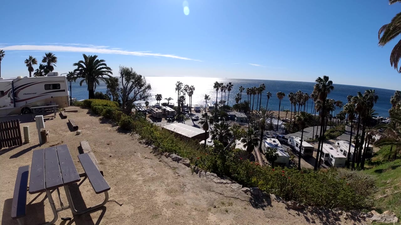 A Malibu hillside view shows a picnic table in the foreground, a camper at left, coastal palm trees, parked cars, bright ocean water, and no visible people.