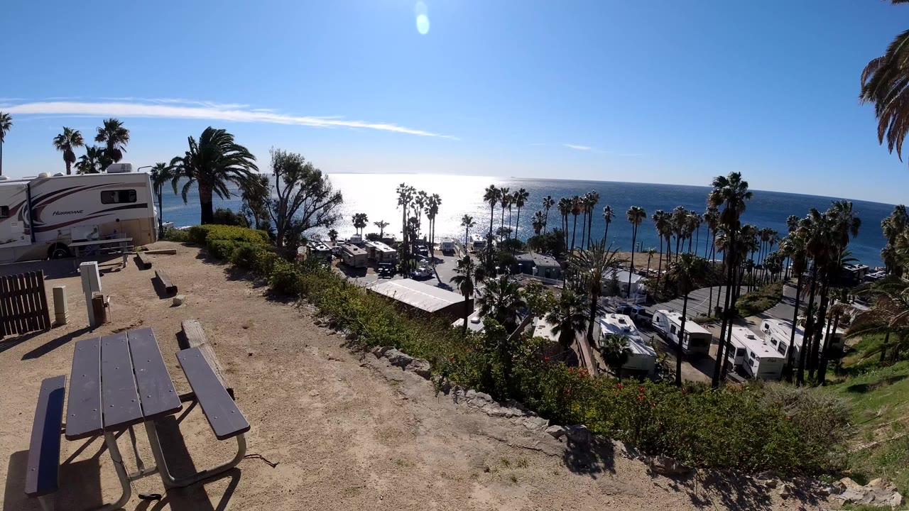 Picnic table and camper above a coastal parking area with palms and ocean.