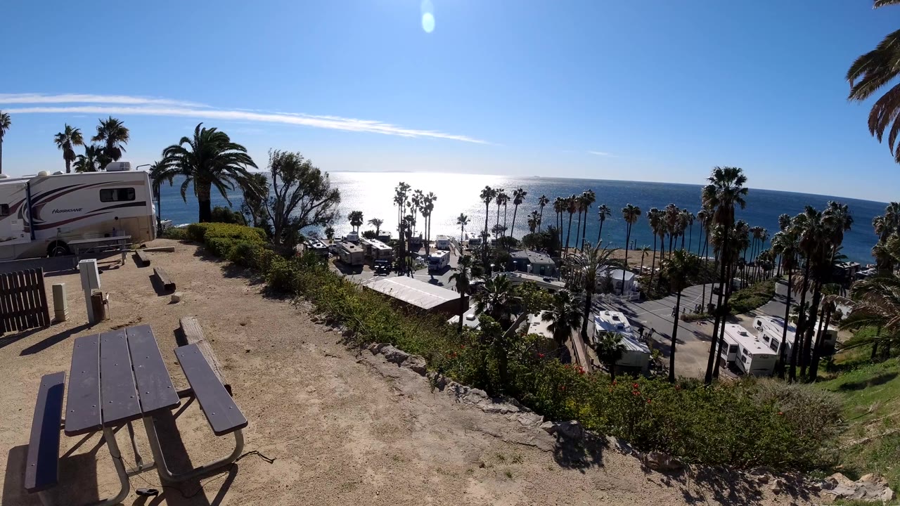 Wide Malibu overlook frame with a picnic table in the dirt foreground, RVs and cars below, palm trees along the coast, blue ocean beyond, and no visible people.