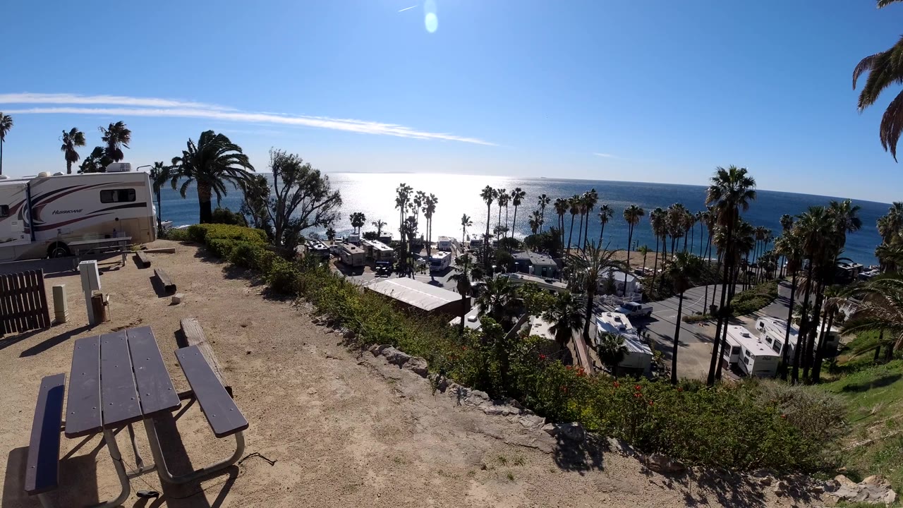 Malibu park overlook frame with dry ground and a picnic table in front, a cluster of RVs and cars below, tall palms by the water, and no visible people.