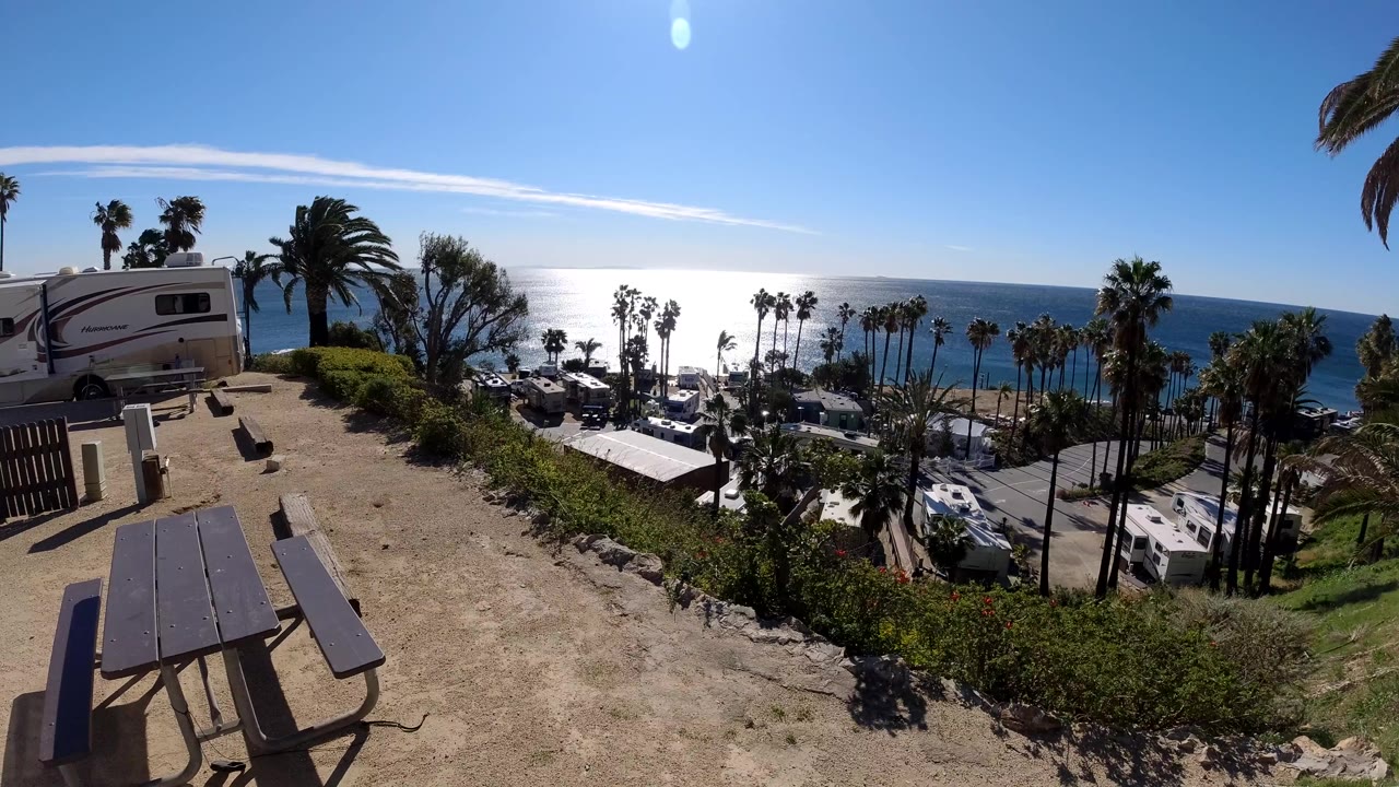 Wide Malibu oceanfront park view with picnic tables in the foreground, RVs below, palm trees along the slope, bright sun on the Pacific, and no visible people.