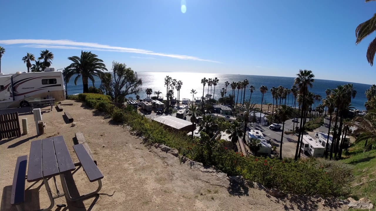 Wide Malibu coastal frame with a picnic table at lower left, an RV at the upper left slope, clustered palms near the water, a bright Pacific horizon, and no visible people.
