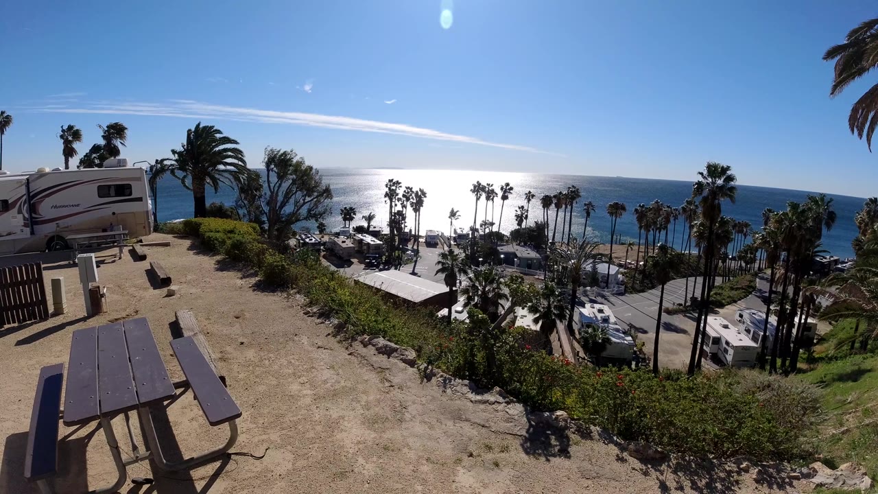 Opening frame of a Malibu, California coastal overlook with picnic tables in the foreground, an RV at left, palm trees below, bright ocean glare, and no visible people.