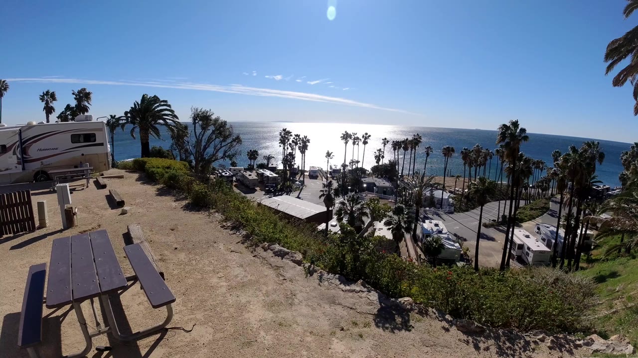 Wide Malibu coastal overlook with a picnic table in the foreground, RVs and palms below, bright ocean glare, blue sky, and no visible people.