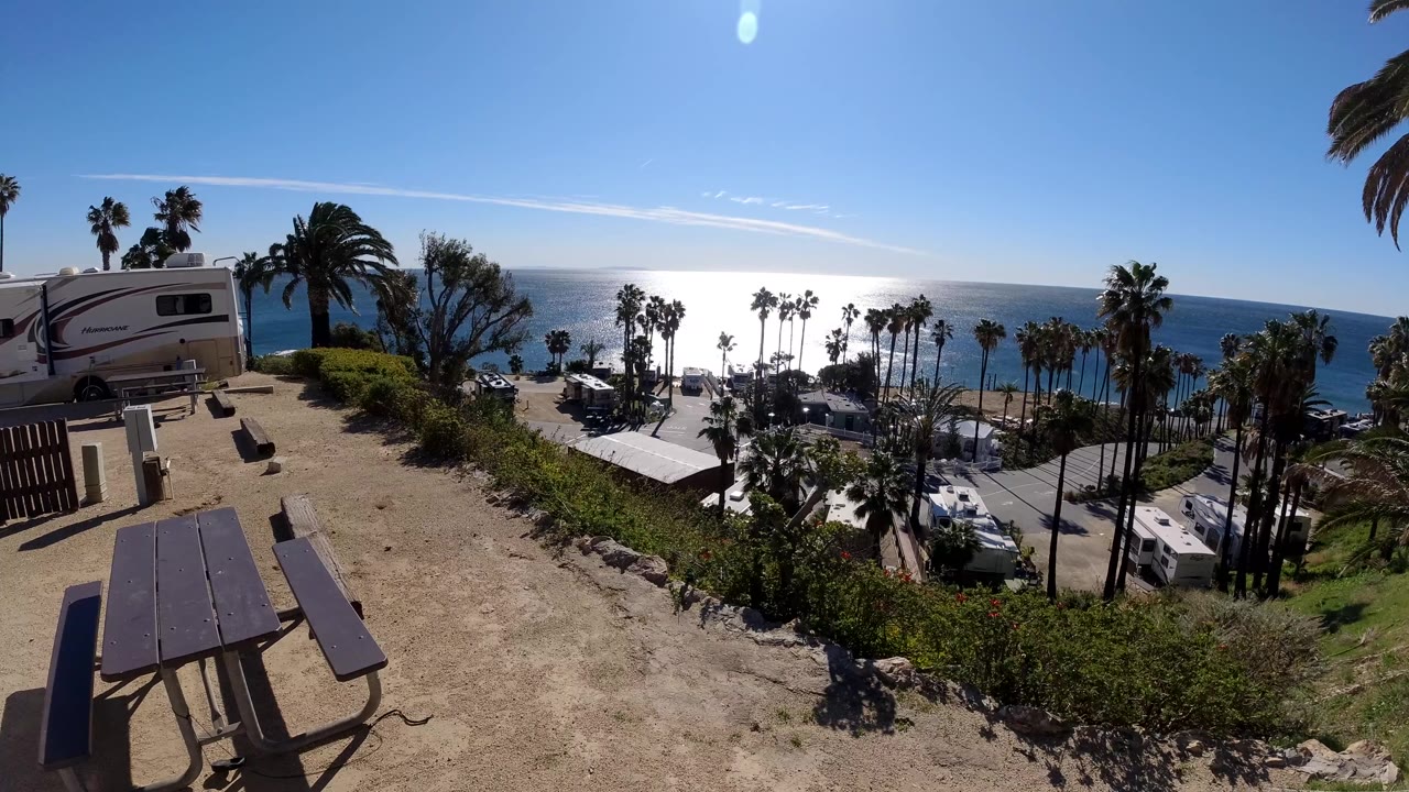 Wide Malibu beachside view at 0:00 with a picnic table in the dirt foreground, RVs near palm trees, bright ocean reflection, and no visible people.