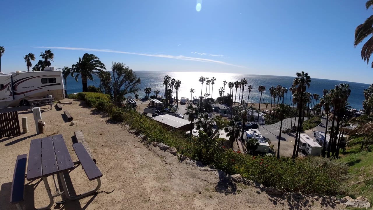 At 0:02, the Malibu overlook frames the left-side RV, foreground dirt path, clustered palms, parked vehicles, bright ocean, and no visible people.