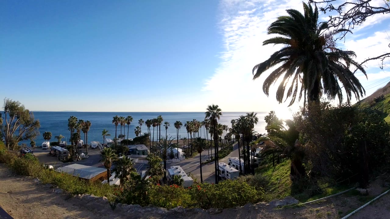 Malibu hillside view with dirt foreground, green brush, palm trees, road traffic, rooftops, blue ocean, sun glare, and no recognizable people visible.