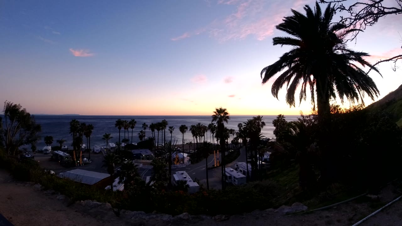 Dark palm trees and hillside foliage frame a Malibu coastal view with rooftops, parked vehicles, ocean waves, and a fading pink sky at the start of the clip.