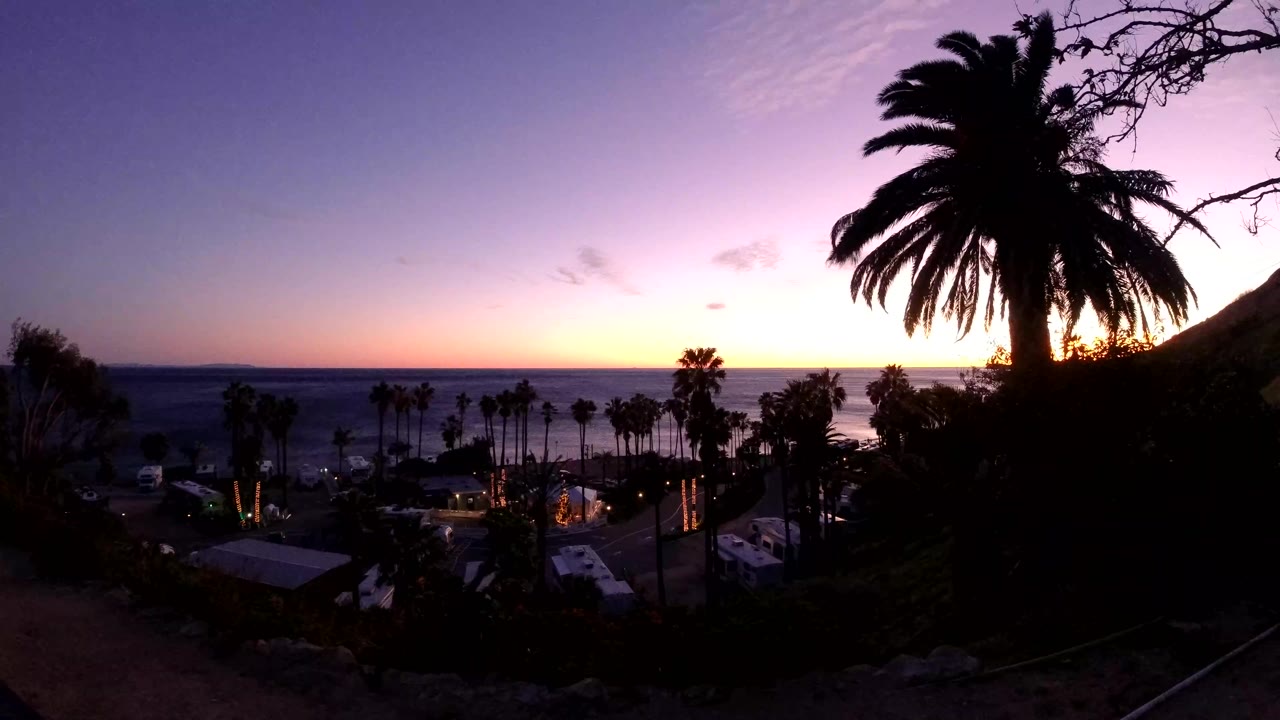 Wide Malibu coastal view with a purple-blue sky, warm light near the sea horizon, silhouetted palms and hillside, and no visible people.