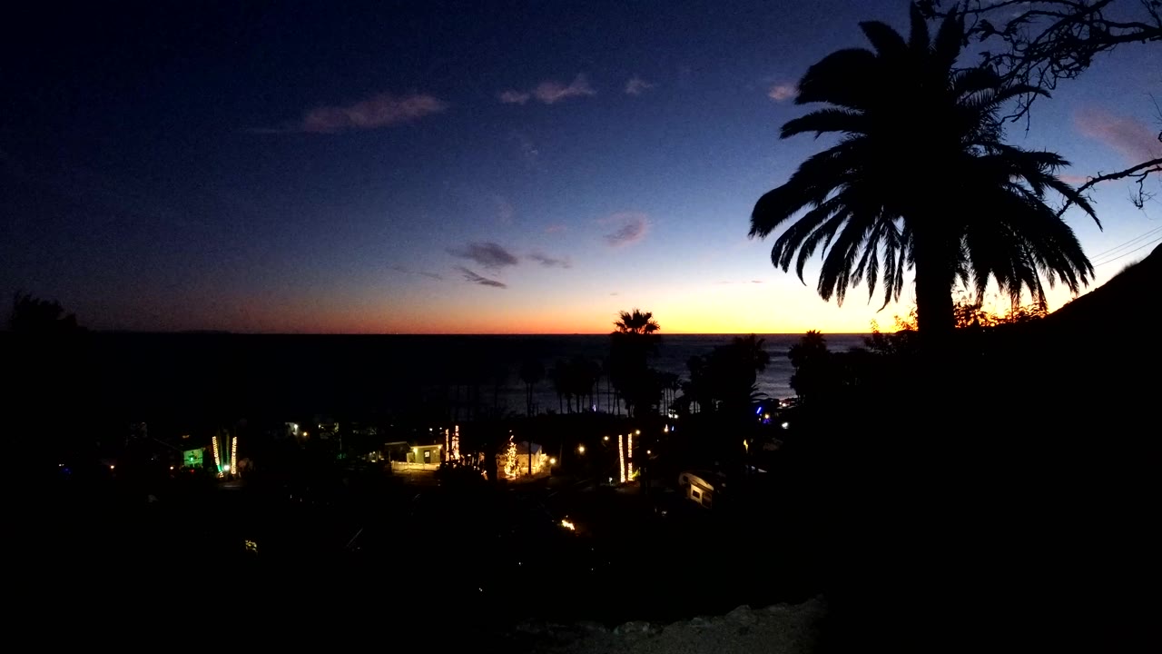 In Malibu, California, the view holds on a broad dusk sky, a large palm silhouette at right, a small palm near the horizon, and scattered lights in the lower frame, with no visible people.