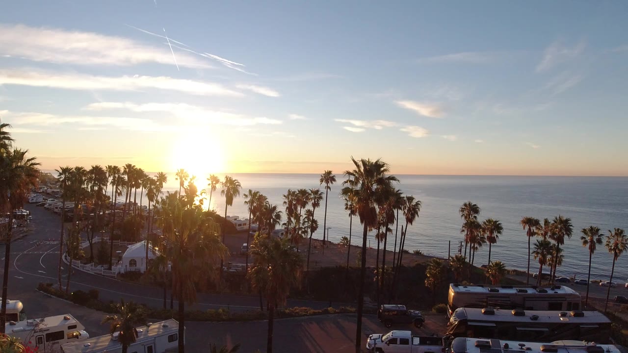 Second frame of the Malibu sunset view, with bright sun flare above the water, dark palm trunks across the beach, a road curve at left, RVs and low buildings in the foreground, and no visible people.