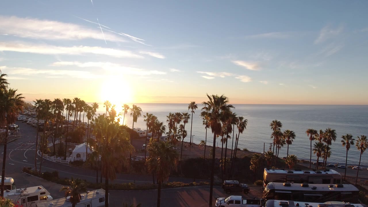 Palm-lined shoreline, bright sunset, coastal road, RVs, and calm ocean water.