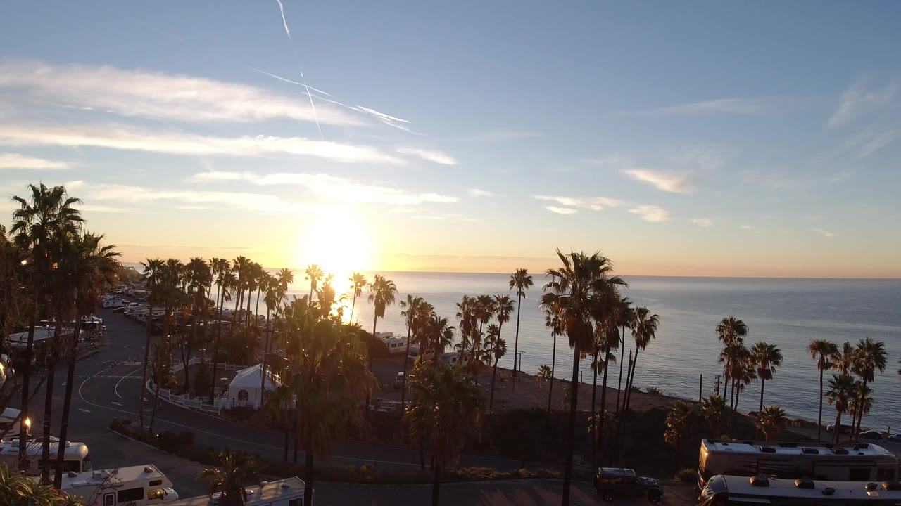 Opening frame of Malibu, California with the sun low over the ocean, palm silhouettes, a curving road, parked vehicles, and no visible people.