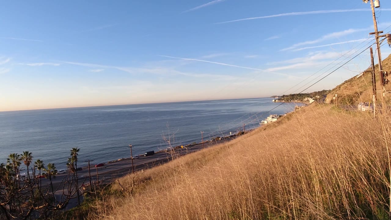 Malibu, California coastal slope with utility poles at the right edge, dry grass across the foreground, ocean beyond, and no visible people.