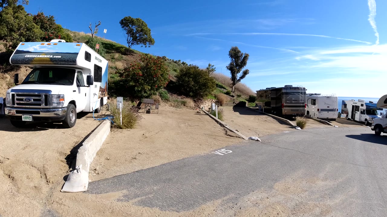 A white Cruise America motorhome sits beside a paved lane in Malibu, California, with other RVs, dirt shoulders, hillside brush, trees, blue sky, and no visible people.