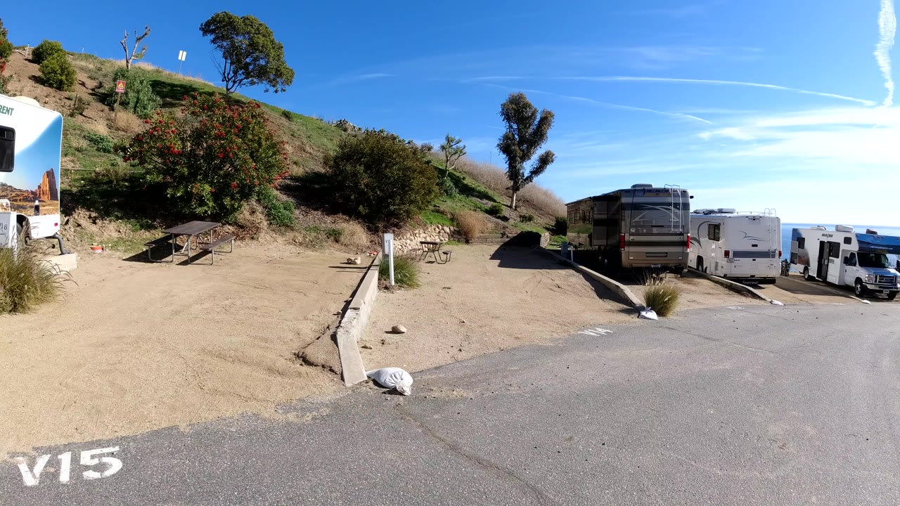 A Malibu, California campground road curves past dirt RV parking, trailers, a picnic table, hillside brush, trees, hard sunlight, and no visible people.