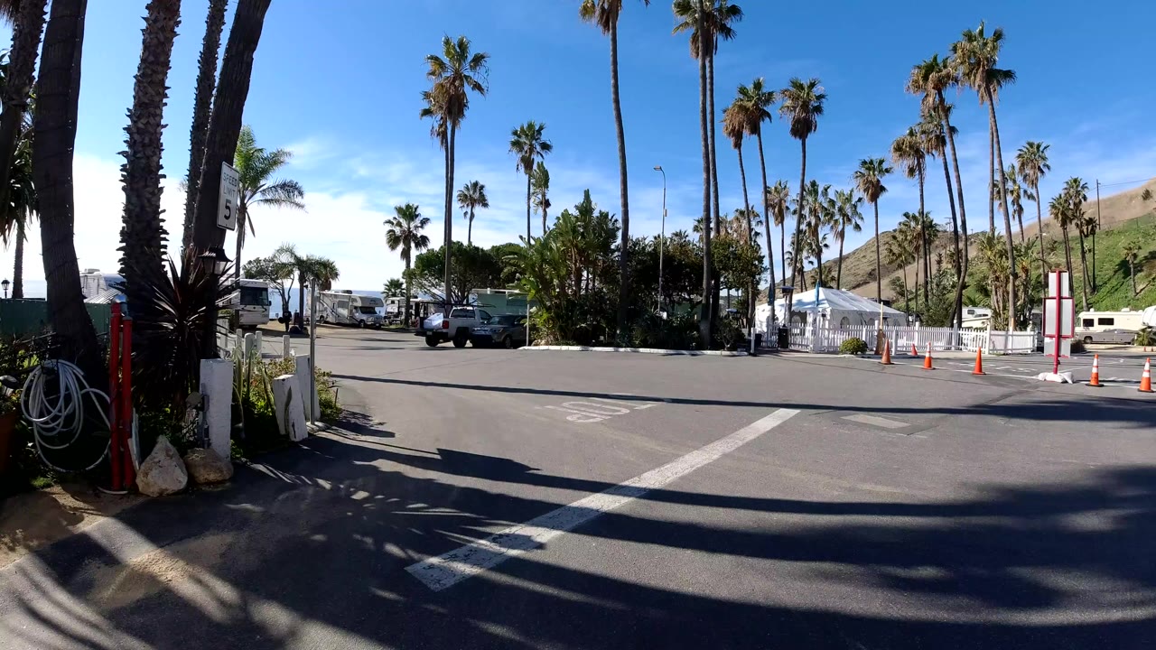 Wide roadside entrance in Malibu with palm trees, orange traffic cones, RVs, parked and moving cars, hills in the background, and no visible people.