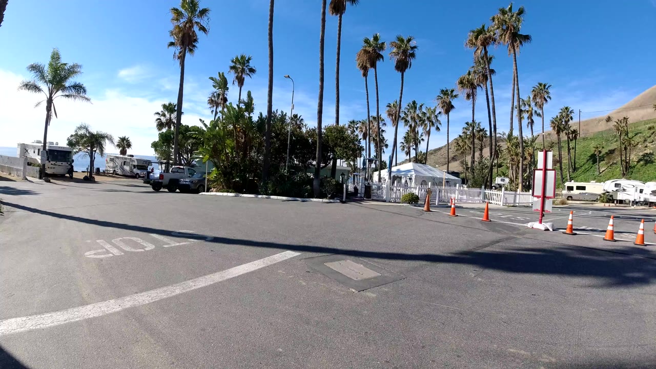 Roadside access road with palm trees, RVs, cones, and hillside.