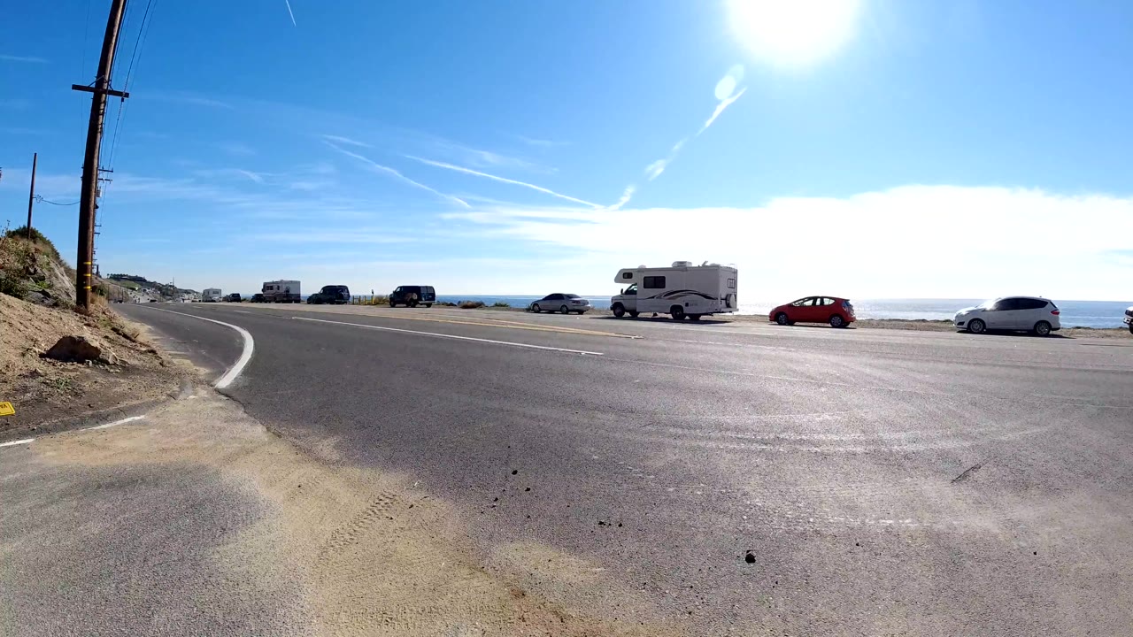 Malibu coastal highway scene with an RV, passing cars, a utility pole, hillside shoulder, ocean visible beyond the road, bright sun, and no visible people.