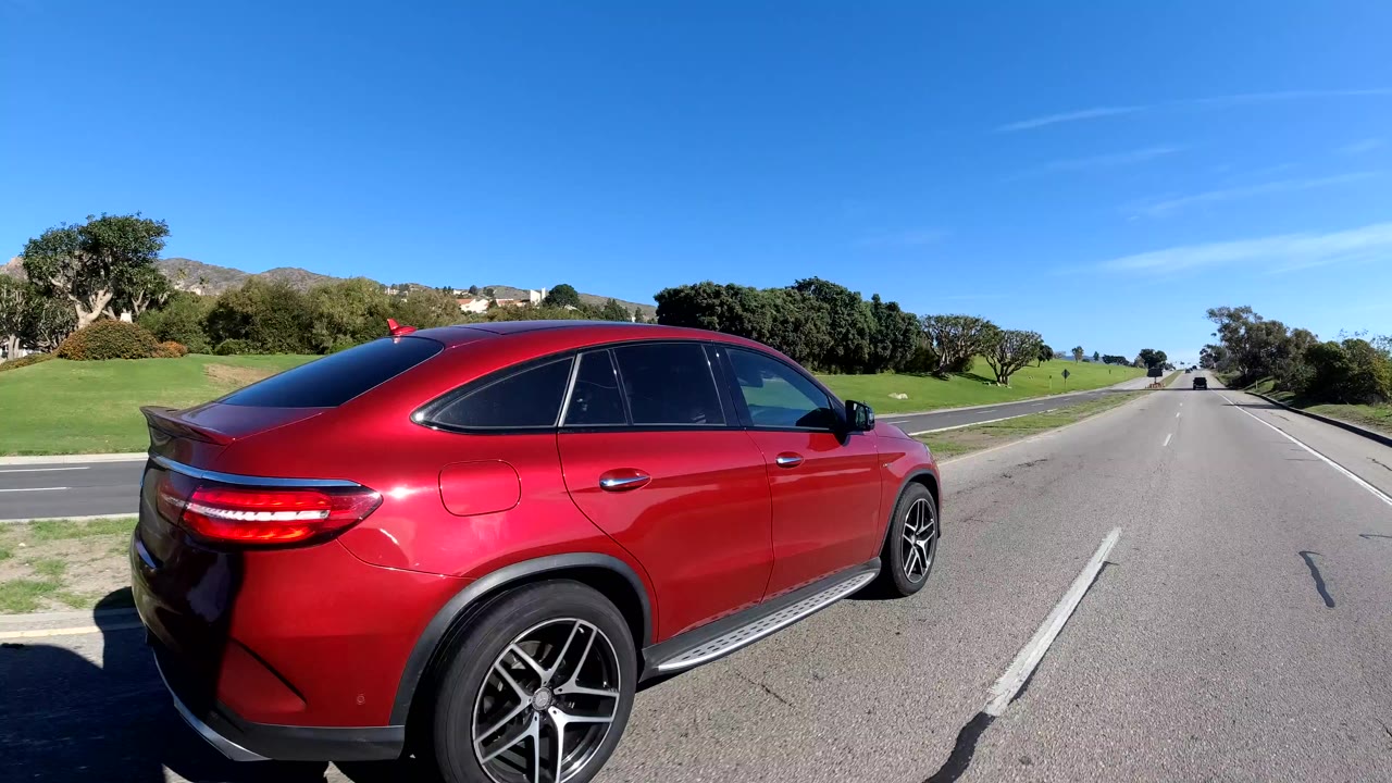 A red SUV fills the left foreground beside a sunny Malibu road, with green roadside grass, distant hills, clear blue sky, and no visible people.